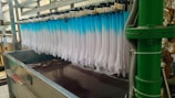 Rows of white fabric are being dyed in a dip-dye method, transitioning into a blue gradient. The setup involves a metal frame holding multiple pieces of fabric over a large container filled with dark liquid. Pipes and industrial equipment are visible around the area.