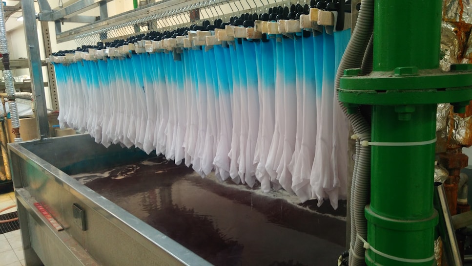 Rows of white fabric are being dyed in a dip-dye method, transitioning into a blue gradient. The setup involves a metal frame holding multiple pieces of fabric over a large container filled with dark liquid. Pipes and industrial equipment are visible around the area.