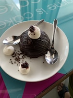 A chocolate dome dessert on a white plate, garnished with whipped cream, chocolate shavings, and a cherry on top. Two spoons are placed on either side, resting on the plate. The background features a glass table with text partially visible.