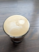 A glass of creamy coffee sits on a wooden surface, with smooth, frothy crema covering the top.