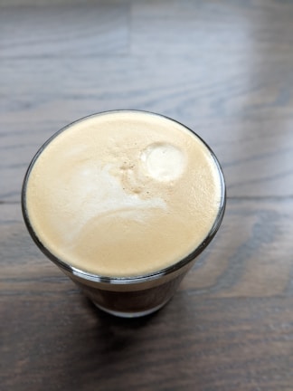 A glass of creamy coffee sits on a wooden surface, with smooth, frothy crema covering the top.