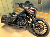 A custom motorcycle with a dark, metallic design featuring shark teeth artwork on the fairing. The bike has a military theme with star insignias and yellow stripes on the side bags. It also boasts large black wheels and a sleek black exhaust system.