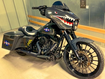 A custom motorcycle with a dark, metallic design featuring shark teeth artwork on the fairing. The bike has a military theme with star insignias and yellow stripes on the side bags. It also boasts large black wheels and a sleek black exhaust system.
