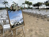 A decorated mirror with floral arrangements on top reflects the surrounding area. White chairs are neatly arranged in rows on a grassy outdoor space, suggesting an event setup. Palm trees and other greenery are visible in the background, under a partly cloudy sky.