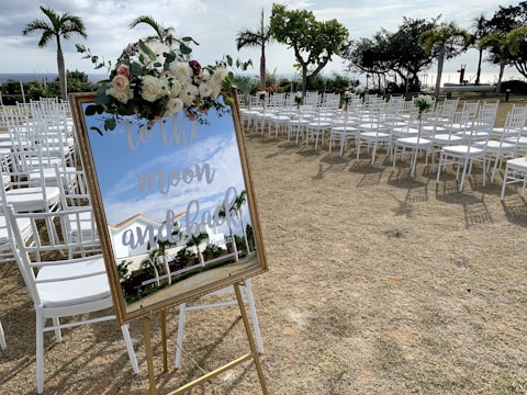 A decorated mirror with floral arrangements on top reflects the surrounding area. White chairs are neatly arranged in rows on a grassy outdoor space, suggesting an event setup. Palm trees and other greenery are visible in the background, under a partly cloudy sky.