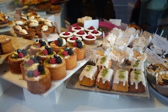 An assortment of pastries and cakes is displayed on platters, featuring a variety of treats topped with fresh berries, icing, and cream. The arrangement includes cupcakes, loaf cakes, and tarts, with labels indicating different flavors.