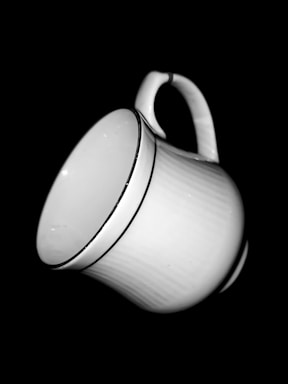 A white ceramic cup with a smooth texture. The cup has a delicate handle and a thin black line near the rim. The background is solid black, making the cup stand out vividly.