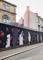 A series of artistic murals painted on a wall, featuring stylized profiles of faces in monochrome, interspersed with vibrant red petals. The wall is set along a street with older buildings in the background, showcasing a blend of architectural and artistic elements.