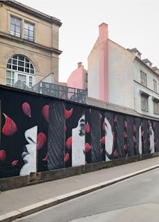 A series of artistic murals painted on a wall, featuring stylized profiles of faces in monochrome, interspersed with vibrant red petals. The wall is set along a street with older buildings in the background, showcasing a blend of architectural and artistic elements.