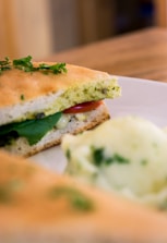 A close-up of a sandwich on a plate, filled with lettuce, a slice of tomato, and melted cheese, garnished with fresh herbs. The sandwich is made with focaccia bread, and there is a scoop of mashed potatoes or similar creamy side dish nearby.