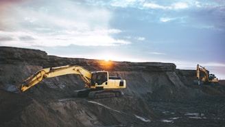 Two large yellow excavators are positioned on a rugged terrain during sunset or sunrise. The machinery is in operation, leveling or excavating the earth with visible tracks on the ground. The sky exhibits a blend of blue, orange, and white hues, suggesting a calm yet industrious environment.