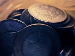 A close-up view of a collection of coins stacked and overlapping each other, highlighting engraved designs on their surfaces with a warm, golden light casting subtle reflections.
