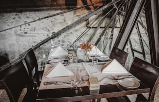 A beautifully set dining table with neatly folded white napkins, empty wine glasses, and floral centerpieces. Dark wooden chairs surround the table, which is set against a backdrop of machinery parts and a stone wall.