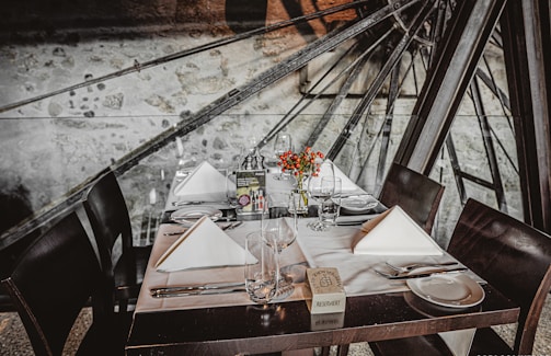A beautifully set dining table with neatly folded white napkins, empty wine glasses, and floral centerpieces. Dark wooden chairs surround the table, which is set against a backdrop of machinery parts and a stone wall.