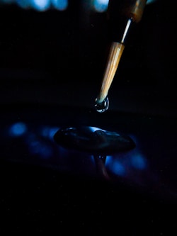 A paintbrush with fine bristles is poised over a surface, releasing a droplet of liquid. The background is dark, creating a dramatic contrast with the illuminated droplet and brush. Reflections can be seen on the surface below.
