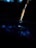 A paintbrush with fine bristles is poised over a surface, releasing a droplet of liquid. The background is dark, creating a dramatic contrast with the illuminated droplet and brush. Reflections can be seen on the surface below.