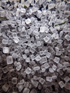 A close-up view of numerous translucent, irregularly shaped crystalline structures resembling small cubes. The crystals appear to be scattered randomly and are closely packed together, showcasing their rough and varied textures. There are hints of green elements subtly visible among the crystals.