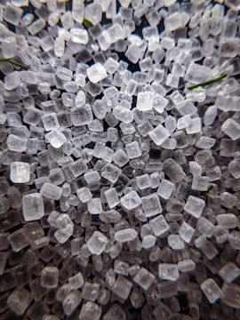 A close-up view of numerous translucent, irregularly shaped crystalline structures resembling small cubes. The crystals appear to be scattered randomly and are closely packed together, showcasing their rough and varied textures. There are hints of green elements subtly visible among the crystals.