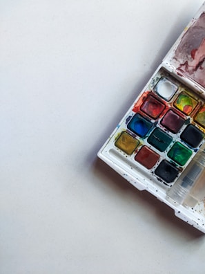 A white background with a rectangular watercolor paint palette positioned in the top right corner. The palette contains multiple compartments filled with various bright colors including red, yellow, green, and blue. Some of the compartments have mixed colors, and there are slight paint splatters around the palette.