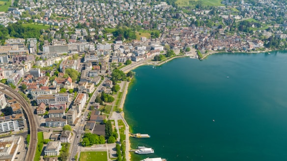 An aerial view of a lakeside town with densely packed buildings, a railway line, and surrounding greenery. The lake has clear, blue-green water, and the shoreline features pathways and a few boats.