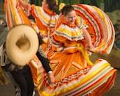 Colorful traditional dancers, wearing vibrant orange dresses adorned with multicolored stripes and ruffles, perform energetically. One dancer holds her dress out gracefully, while another wears a wide-brimmed hat and detailed black attire.
