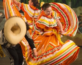 Colorful traditional dancers, wearing vibrant orange dresses adorned with multicolored stripes and ruffles, perform energetically. One dancer holds her dress out gracefully, while another wears a wide-brimmed hat and detailed black attire.