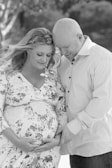 A tender moment between a couple as they embrace, with the woman gently cradling her pregnant belly. Both are smiling softly, sharing an intimate and affectionate connection. The outdoor setting in black and white adds a timeless and serene feel.