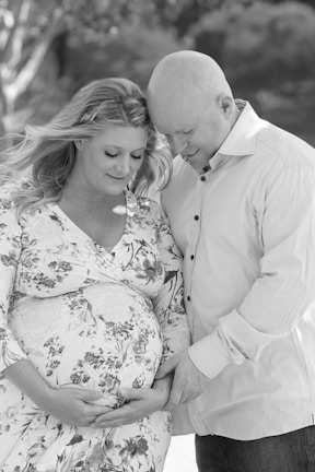 A tender moment between a couple as they embrace, with the woman gently cradling her pregnant belly. Both are smiling softly, sharing an intimate and affectionate connection. The outdoor setting in black and white adds a timeless and serene feel.