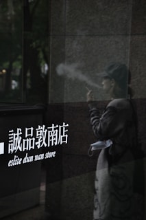 A person is seen smoking beside a reflective surface with the text 'eslite dun nan store' displayed. The reflection shows the person wearing a cap and a casual outfit. A mask hangs from the wrist or arm. The lighting is dim, creating a contemplative or subdued atmosphere.