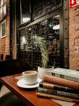 A cozy setting features a wooden table with a white coffee cup on a saucer, several books stacked beside it, and a potted plant in the background. The backdrop includes a brick wall and large, intricately designed iron-framed window, offering a glimpse of a dimly lit interior with warm lighting.