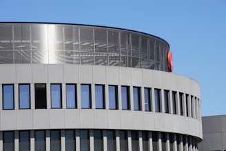 A modern architectural structure with a cylindrical design features large glass windows and a sleek concrete facade. The building's upper section shows reflections and linear patterns inside. An orange and red logo is visible on the curved surface.