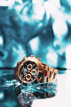 A luxurious gold wristwatch with an intricate design and multiple dials is reflecting against a smooth glass surface. The background is a blurred mix of vibrant blue hues, adding depth and a sense of elegance to the composition.