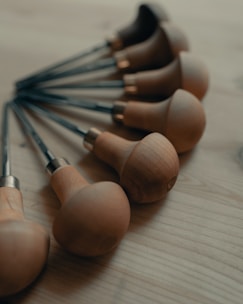 A set of woodworking carving tools with wooden handles neatly arranged on a wooden surface. Each tool has a smooth, rounded wooden grip and a metal shaft extending outward.