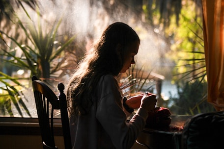 A woman journaling by a sunlit window, surrounded by plants, reflecting self-discovery.