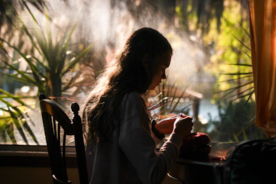 A serene morning scene with a woman journaling by a sunlit window, surrounded by soft greenery.