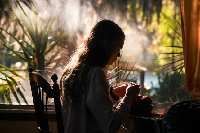 A person is sitting by a window, focused on a task with their hands. Sunlight streams through the window, illuminating their hair and creating a serene atmosphere. Lush greenery is visible outside the window.
