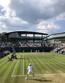 A tennis match is taking place on a well-maintained grass court surrounded by a large audience seated in the stands. The scene shows a player in a white outfit positioned near the net while the other player is ready to serve. A modern stadium building with large windows and a roof overhang is visible in the background under a partly cloudy sky.