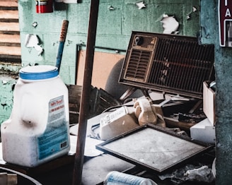 An old, dusty workspace filled with various objects including a large white plastic container, a vintage rotary dial telephone, a broken cassette player, a wall-mounted air conditioning unit, and scattered debris. The peeling paint on the walls indicates neglect and decay.