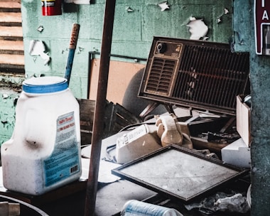 An old, dusty workspace filled with various objects including a large white plastic container, a vintage rotary dial telephone, a broken cassette player, a wall-mounted air conditioning unit, and scattered debris. The peeling paint on the walls indicates neglect and decay.