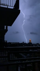 A dramatic scene of a lightning bolt striking against a cloudy, night-time sky. A silhouetted view of building balconies frames the left side, while a distant illuminated tower stands out amidst the urban landscape.
