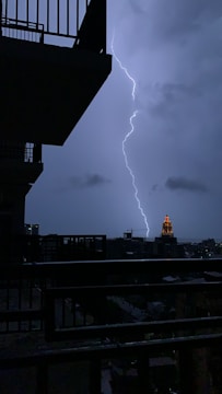 A dramatic scene of a lightning bolt striking against a cloudy, night-time sky. A silhouetted view of building balconies frames the left side, while a distant illuminated tower stands out amidst the urban landscape.