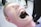 A dental mannequin head with an open mouth showing detailed teeth. The mannequin has a smooth, human-like face with closed eyes and is positioned on a plain surface. In the background, there are blurred figures and furniture, suggesting an indoor setting possibly related to dentistry or medical education.