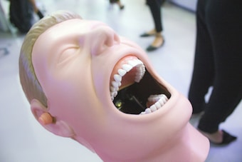 A dental mannequin head with an open mouth showing detailed teeth. The mannequin has a smooth, human-like face with closed eyes and is positioned on a plain surface. In the background, there are blurred figures and furniture, suggesting an indoor setting possibly related to dentistry or medical education.