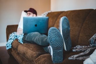 A person wearing casual clothing and a cap is reclining on a brown couch while using a laptop. The laptop displays a recognizable logo on its lid. One foot is visible with its sneaker sole facing outward. A patterned blanket or throw is draped on the couch.