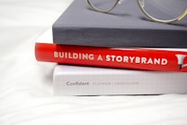 A stack of three books is neatly arranged, with the top book covered in a plain gray hardcover. Below it is a bright red hardcover book titled 'Building a StoryBrand', followed by a white grid-patterned hardcover labeled 'Confidant Planner / Hardcover'. A pair of glasses with round lenses rests on top of the stack, adding a scholarly touch.
