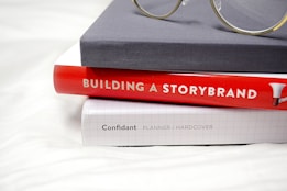 A stack of three books is neatly arranged, with the top book covered in a plain gray hardcover. Below it is a bright red hardcover book titled 'Building a StoryBrand', followed by a white grid-patterned hardcover labeled 'Confidant Planner / Hardcover'. A pair of glasses with round lenses rests on top of the stack, adding a scholarly touch.