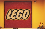 A square display of the LEGO logo composed of numerous small red, white, and yellow pieces on a red background, mounted on a yellow wall. In the foreground, there are various LEGO models and a security camera visible in the upper right corner.