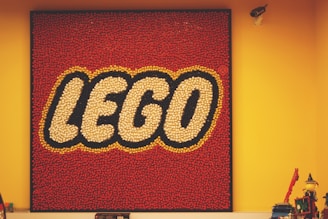A square display of the LEGO logo composed of numerous small red, white, and yellow pieces on a red background, mounted on a yellow wall. In the foreground, there are various LEGO models and a security camera visible in the upper right corner.