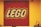 A square display of the LEGO logo composed of numerous small red, white, and yellow pieces on a red background, mounted on a yellow wall. In the foreground, there are various LEGO models and a security camera visible in the upper right corner.