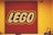 A square display of the LEGO logo composed of numerous small red, white, and yellow pieces on a red background, mounted on a yellow wall. In the foreground, there are various LEGO models and a security camera visible in the upper right corner.